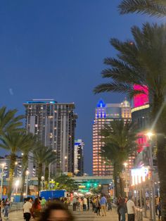 many people are walking on the sidewalk in front of palm trees and tall buildings at night