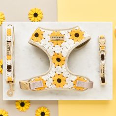 A **Sunflower Patch Adjustable Neck Harness, Lead & Collar Bundle** by **Cocopup London**, featuring a vibrant sunflower pattern in yellow and white, is displayed on a white marble surface. Surrounding the harness are the matching leash and collar, perfect for stylish dog walks. Additional small artificial sunflowers are placed around the items for decoration. Personalised Jute Bags, Sunflower Patch, Sunflower Crochet, Travel Dog Bowl, Dog Walking Bag, Treat Pouch, Walking Bag, Yellow Sunflowers, Dog Walks