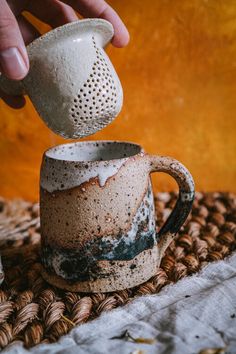 a person is pouring something into a coffee mug on a woven tablecloth with other items nearby