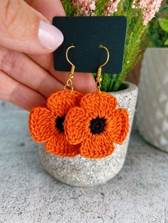 an orange crocheted flower earrings is being held in front of a pot with pink flowers