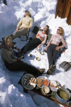 four people are lounging in the snow with food and drinks on their lap tops