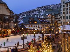 people skating on an ice rink at night in front of buildings with lit christmas trees