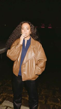 a woman standing in the dark with her hand on her mouth and wearing a brown leather jacket