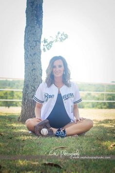 a woman sitting on the ground in front of a tree and wearing a baseball uniform