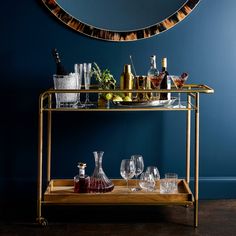 a gold bar cart with glasses and bottles on it in front of a blue wall