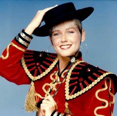 a woman in a red jacket and black hat smiles at the camera with her hand on her head