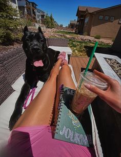 a person sitting on a deck next to a black dog with a drink in their hand