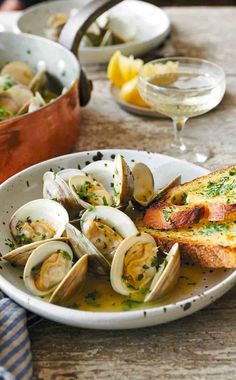 clams and bread in a white bowl on a wooden table next to wine glasses