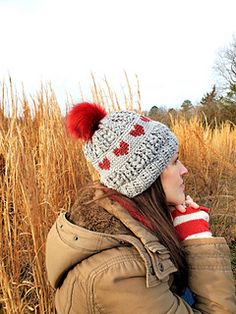 a woman sitting in tall grass wearing a knitted hat with hearts on it and a red pom - pom