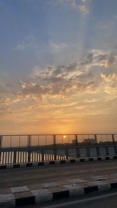 the sun is setting behind a fence and some cars are driving on the road beside it