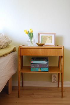 a small wooden table with some books on it next to a bed and flowers in a vase