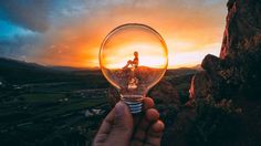 a person holding up a light bulb in front of a mountain range with the sun setting behind them