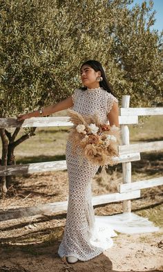 a woman standing in front of an olive tree wearing a white dress and holding a bouquet