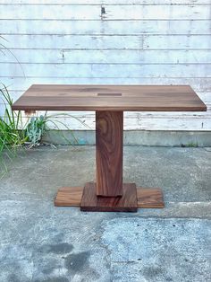 a wooden table sitting on top of a cement floor next to a white wall and green plants