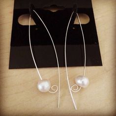 two white pearls are hanging from long silver earwires on a black piece of paper