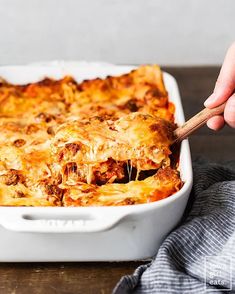 a hand holding a wooden spoon over a casserole dish filled with meat and cheese