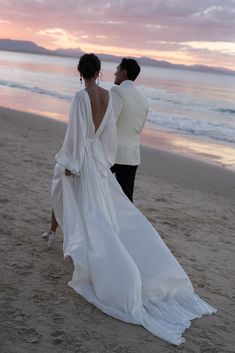 a bride and groom walking on the beach at sunset with their back to the camera