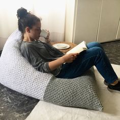 a woman sitting on a bean bag chair reading a book and holding a coffee cup