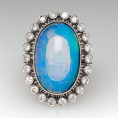 This gorgeous ring is centered with one (1) oval cabochon cut natural crystal opal and set into a bezel setting. The opal is bordered with twenty-two (22), bezel set, round brilliant cut diamonds, The top face of the shank is accented with three (3), bead set, round brilliant cut diamonds on each side. The gemstones are bordered with milgrain edging. The ring measures 29.1mm at the top, rises 10.6mm above the finger, tapering to 1.8mm wide and 1.1mm thick at the base of the shank. This ring is currently a size 6. Oval Cabochon Opal Ring Fine Jewelry, Fine Jewelry Oval Cabochon Opal Ring, Oval Cabochon Opal Ring In Fine Jewelry, Opal Diamond Ring Oval Cabochon, Diamond Opal Ring Oval Cabochon, Oval Opal Ring With Bezel Setting For Formal Occasions, Oval Cabochon Diamond Opal Ring, Luxury Oval Opal Ring With Bezel Setting, Elegant Opal Ring With Oval Cabochon Bezel Setting