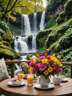 a table with flowers and fruit on it in front of a waterfall, surrounded by greenery