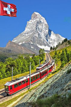 a red train traveling past a tall mountain covered in snow next to green grass and trees