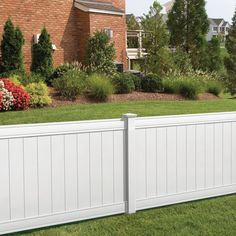 a white vinyl fence in front of a brick building with flowers and shrubs around it