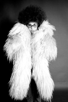 an old photo of a woman with glasses and a fur coat over her shoulders, standing in front of a black background