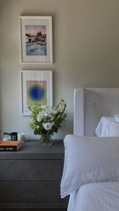 a white bed sitting next to a nightstand with flowers on top of it and two pictures above it