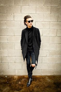 a man in black jacket and sunglasses standing next to a brick wall with his hands in his pockets