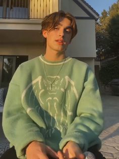 a young man sitting on the ground in front of a house wearing a green sweater