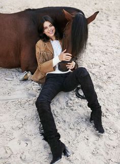 a woman is sitting in the sand next to a horse and posing for a photo