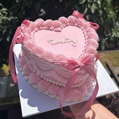 a heart shaped cake with pink icing and ribbon on top, being held by someone