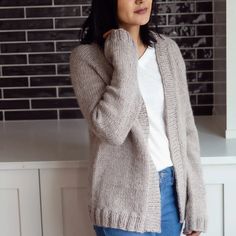 a woman standing in front of a window wearing a gray cardigan sweater and jeans