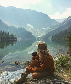 a woman sitting on the edge of a lake holding a coffee cup in her hand