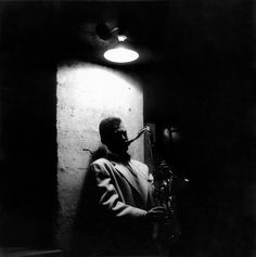 black and white photograph of man playing saxophone in dark room with light coming from ceiling