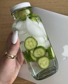 a person holding a glass jar filled with cucumbers and ice on top of a laptop