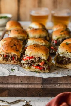 mini sandwiches with meat, peppers and cheese on a cutting board next to glasses of beer