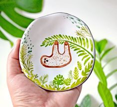 a hand holding a small plate with a sloth on it's side and green leaves around the edge