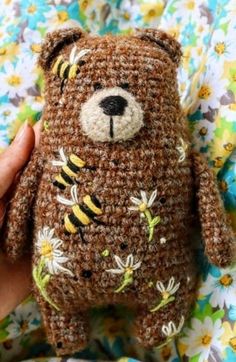 a hand holding a brown crocheted teddy bear with bees on it's chest