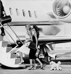 two women are walking towards an airplane with their small dog on the ground next to them