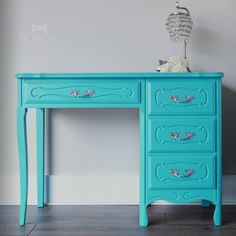 a blue painted desk with flowers on top and a vase in the corner next to it