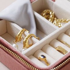 an open pink box with gold rings and jewelry in it on a table next to a white cloth