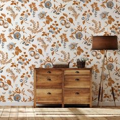 a wooden dresser sitting next to a lamp on top of a hard wood floor in front of a floral wallpaper