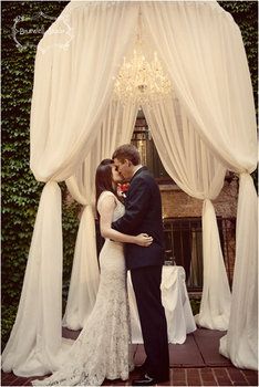 a man and woman are kissing under a wedding arch with white drapes on it