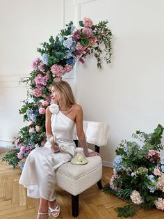 a woman is sitting on a chair with flowers around her and holding a cup in her hand