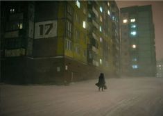 a woman walking down a snow covered street in the middle of winter with tall buildings behind her