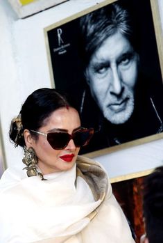 a woman wearing sunglasses and a white coat in front of a poster with a man's face on it