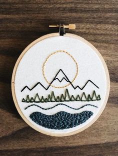 an embroidery project with mountains and trees on the hoop hanging from a wooden table top