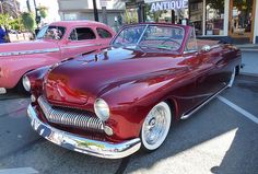 an old red car parked in a parking lot next to another classic car on the street