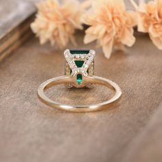 an emerald and diamond ring sitting on top of a wooden table with flowers in the background